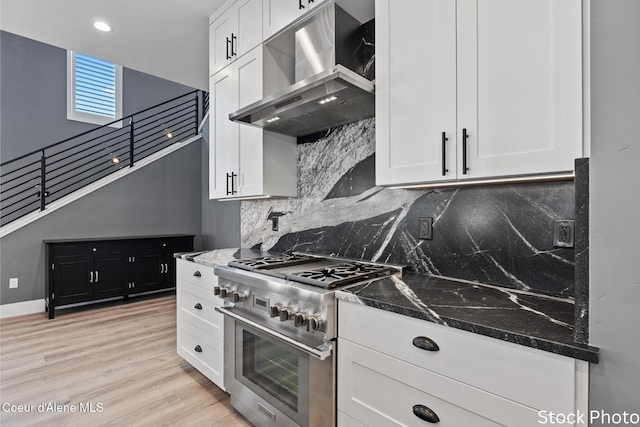 kitchen featuring wall chimney range hood, dark stone countertops, stainless steel stove, and white cabinets