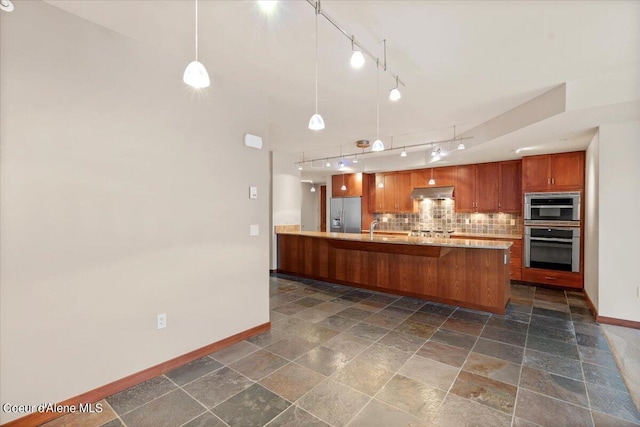 kitchen with under cabinet range hood, hanging light fixtures, appliances with stainless steel finishes, brown cabinets, and tasteful backsplash