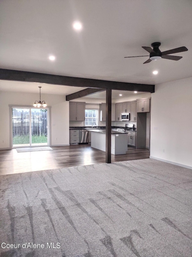 unfurnished living room with sink, dark carpet, ceiling fan with notable chandelier, and beam ceiling