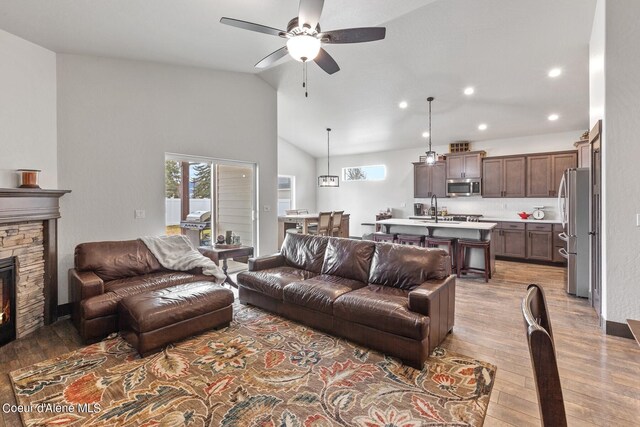 living room featuring light wood-type flooring, ceiling fan, a stone fireplace, sink, and high vaulted ceiling