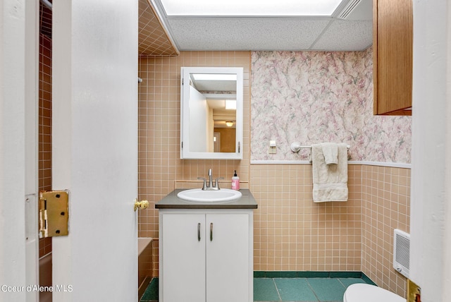 bathroom with wallpapered walls, a drop ceiling, wainscoting, vanity, and tile walls