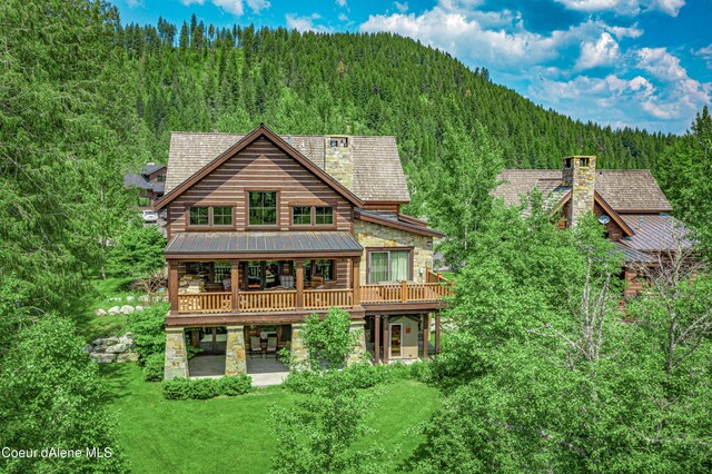 back of house featuring stone siding, a wooded view, a yard, and a patio area