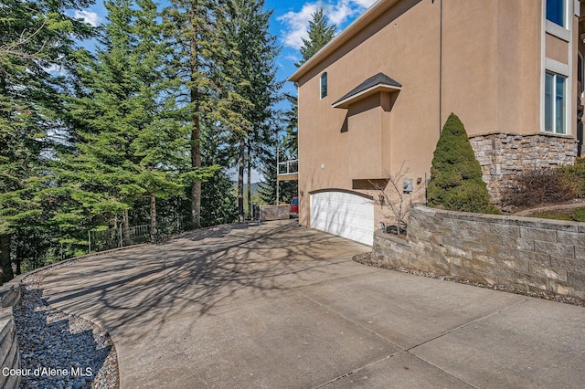 view of side of home with stone siding, fence, an attached garage, concrete driveway, and stucco siding