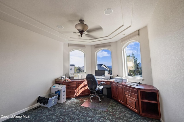 office space with a tray ceiling, a textured wall, baseboards, dark carpet, and a ceiling fan