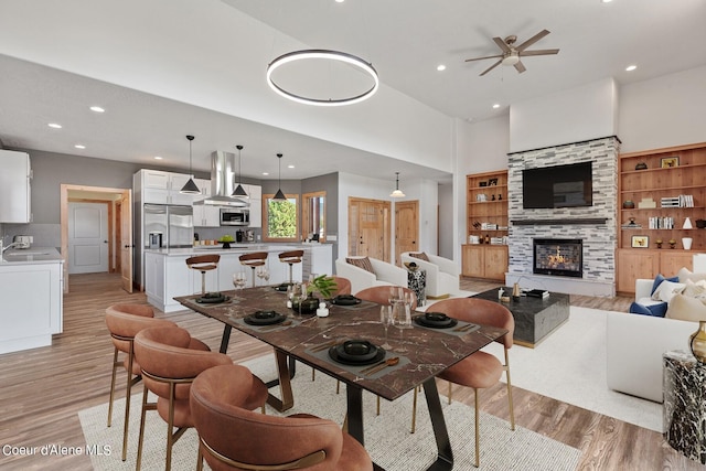 dining room with ceiling fan, built in features, a fireplace, a towering ceiling, and light hardwood / wood-style floors