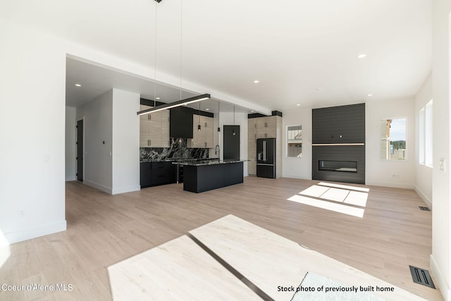 kitchen with light wood-type flooring, black fridge, backsplash, a center island with sink, and decorative light fixtures