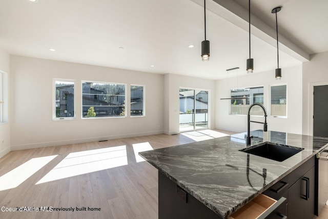 kitchen with dark stone counters, light hardwood / wood-style floors, sink, an island with sink, and hanging light fixtures