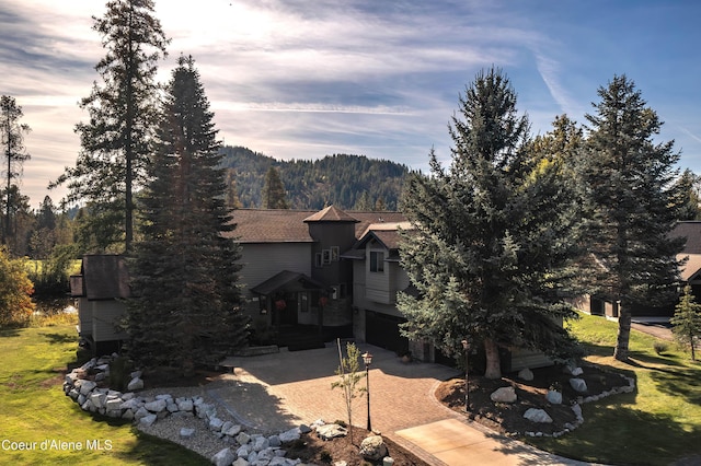 back of house featuring an attached garage, a shingled roof, a forest view, and decorative driveway