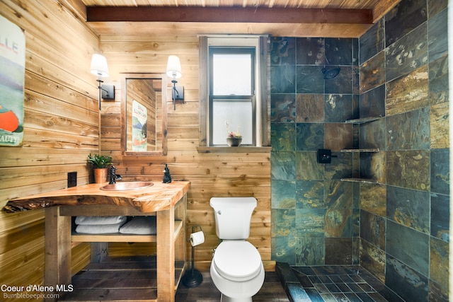 bathroom featuring vanity, beam ceiling, wooden walls, a tile shower, and toilet