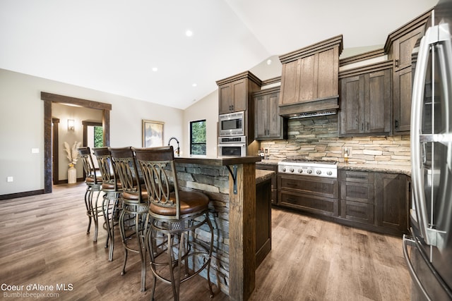 kitchen with an island with sink, light wood-type flooring, dark brown cabinetry, stainless steel appliances, and vaulted ceiling