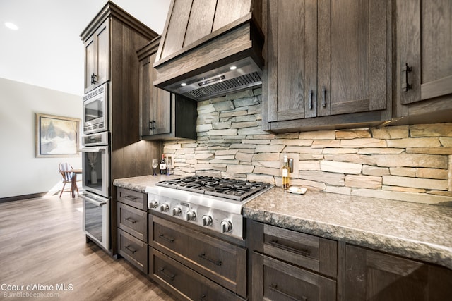 kitchen with light hardwood / wood-style floors, dark brown cabinets, custom exhaust hood, backsplash, and appliances with stainless steel finishes