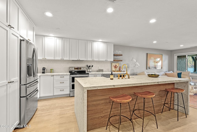 kitchen featuring a kitchen island with sink, white cabinetry, appliances with stainless steel finishes, light wood-type flooring, and a kitchen bar