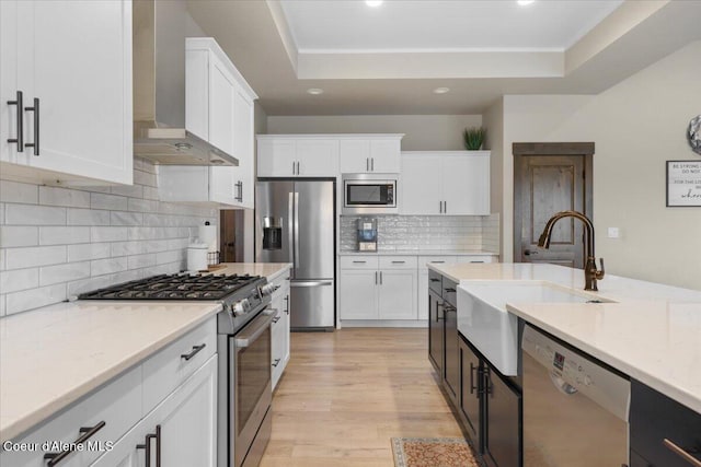 kitchen featuring wall chimney exhaust hood, light hardwood / wood-style flooring, white cabinets, a raised ceiling, and appliances with stainless steel finishes