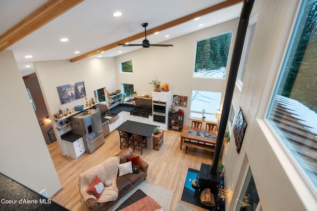 living room featuring ceiling fan, high vaulted ceiling, beamed ceiling, light hardwood / wood-style floors, and a wood stove