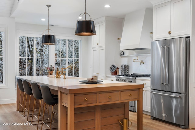 kitchen with high quality appliances, a center island with sink, custom range hood, decorative light fixtures, and white cabinetry