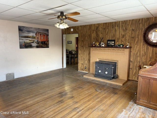 unfurnished living room with a paneled ceiling, a wood stove, wooden walls, hardwood / wood-style flooring, and ceiling fan