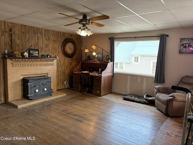 living room featuring a drop ceiling, ceiling fan, wooden walls, hardwood / wood-style flooring, and a wood stove