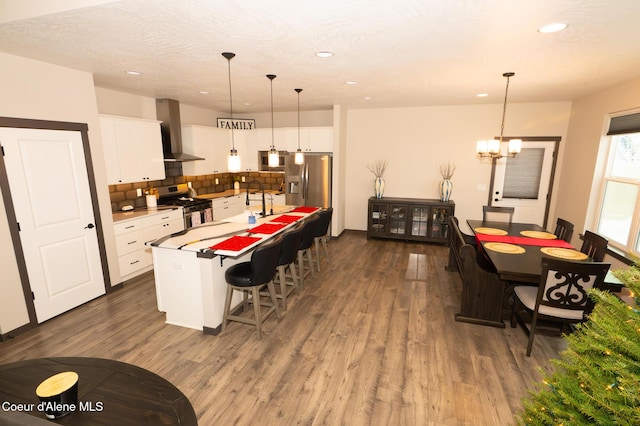 kitchen featuring a kitchen island with sink, white cabinets, wall chimney exhaust hood, appliances with stainless steel finishes, and decorative light fixtures