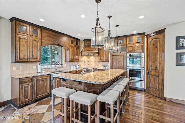 kitchen with tasteful backsplash, custom range hood, stainless steel double oven, dark hardwood / wood-style floors, and an island with sink