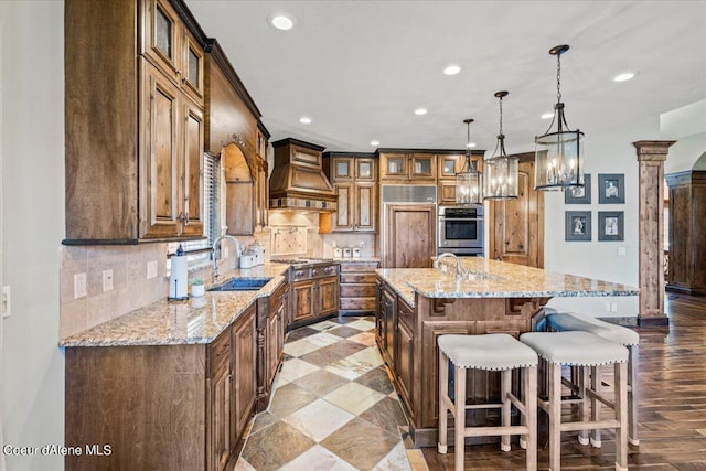 kitchen with a center island with sink, sink, light wood-type flooring, custom range hood, and decorative columns