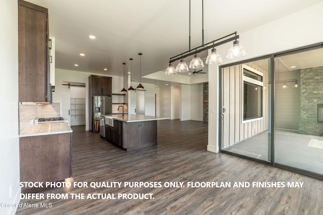 kitchen with light stone counters, dark brown cabinets, dark wood-type flooring, pendant lighting, and an island with sink