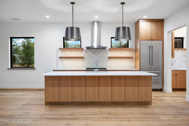 kitchen featuring decorative light fixtures, stainless steel built in fridge, wall chimney range hood, and an island with sink