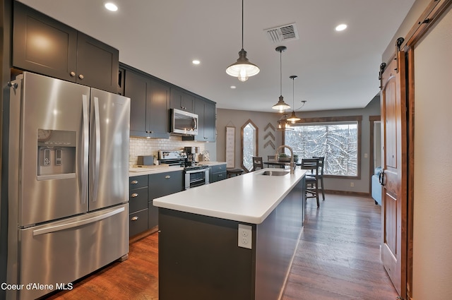 kitchen featuring appliances with stainless steel finishes, a kitchen island with sink, sink, a barn door, and hanging light fixtures