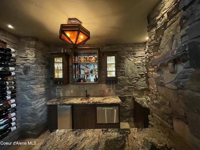 bar with a textured ceiling, dark brown cabinetry, stainless steel dishwasher, and dark stone counters