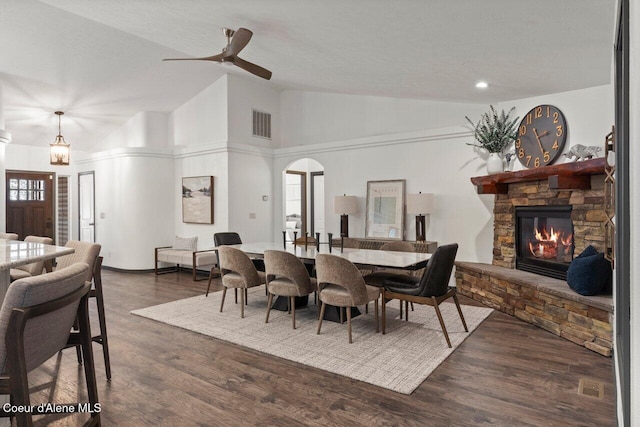 dining room featuring a stone fireplace, ceiling fan, high vaulted ceiling, and dark wood-type flooring