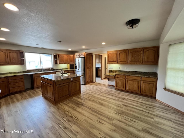 kitchen featuring an island with sink, dark stone counters, stainless steel refrigerator with ice dispenser, sink, and light hardwood / wood-style flooring