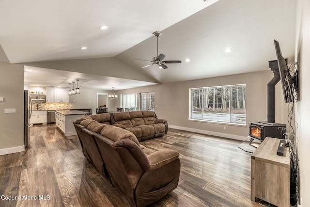living area with dark wood finished floors, vaulted ceiling, ceiling fan with notable chandelier, baseboards, and a wood stove