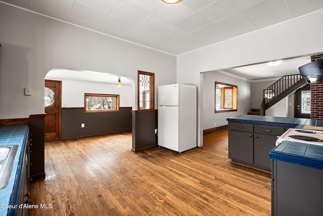 kitchen with light wood finished floors, tile counters, arched walkways, freestanding refrigerator, and gray cabinetry