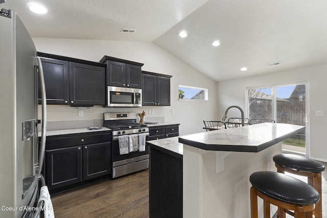 kitchen featuring dark wood-type flooring, a textured ceiling, a kitchen bar, a center island with sink, and appliances with stainless steel finishes