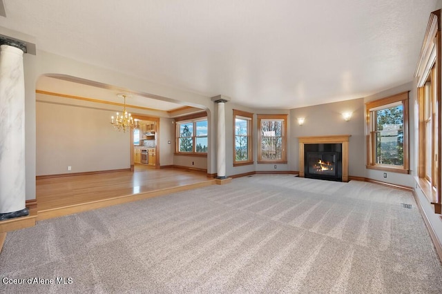 unfurnished living room featuring a glass covered fireplace, a chandelier, baseboards, carpet flooring, and ornate columns