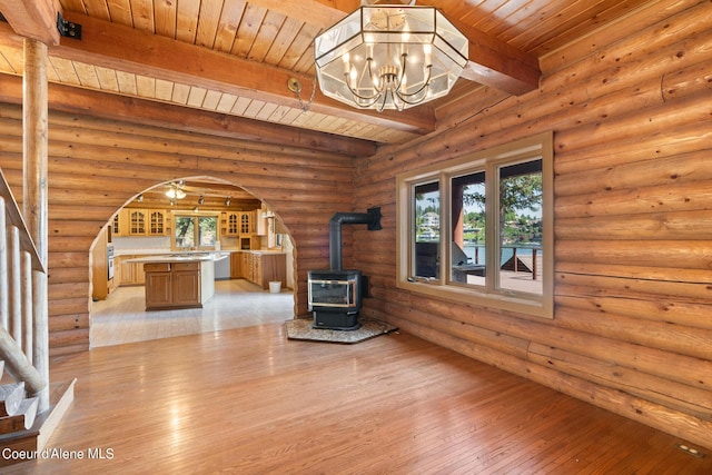 unfurnished living room with a chandelier, light wood-type flooring, beamed ceiling, a wood stove, and rustic walls