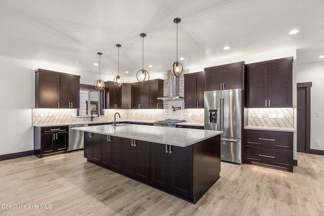 kitchen featuring hanging light fixtures, a kitchen island with sink, appliances with stainless steel finishes, light wood-type flooring, and wall chimney exhaust hood