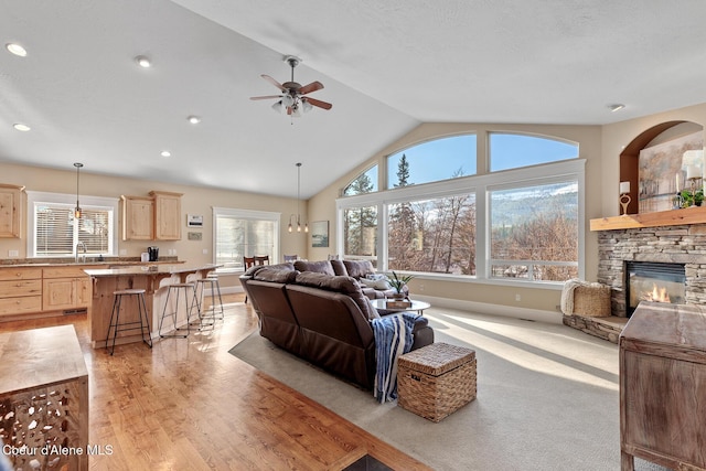 living room featuring vaulted ceiling, light hardwood / wood-style floors, sink, a stone fireplace, and ceiling fan with notable chandelier