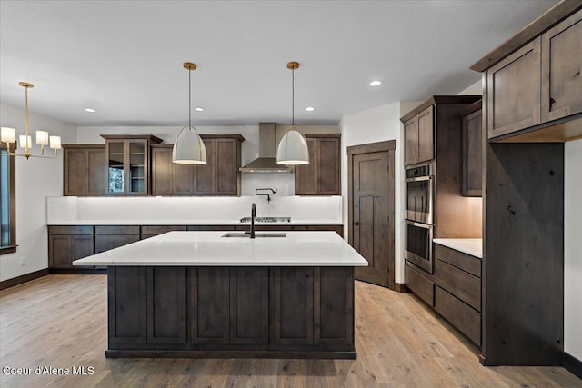 kitchen with wall chimney exhaust hood, decorative light fixtures, double oven, an island with sink, and light hardwood / wood-style floors