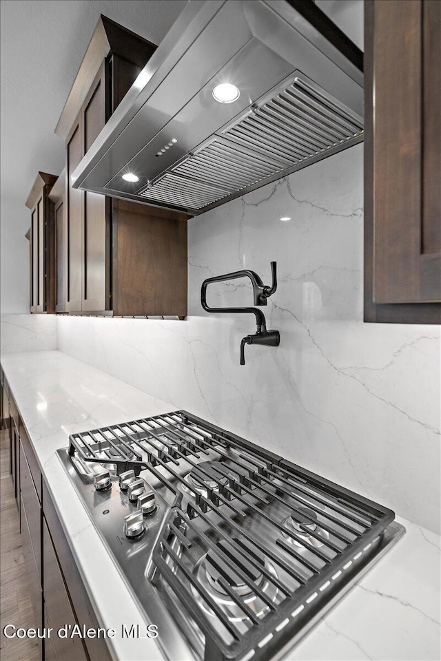 kitchen with dark brown cabinets, backsplash, light stone counters, and wall chimney range hood