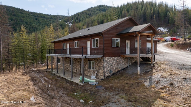 view of property exterior with a patio, a view of trees, stone siding, and a shingled roof