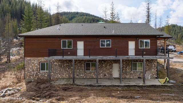 back of property with stone siding, a patio, roof with shingles, and a wooded view