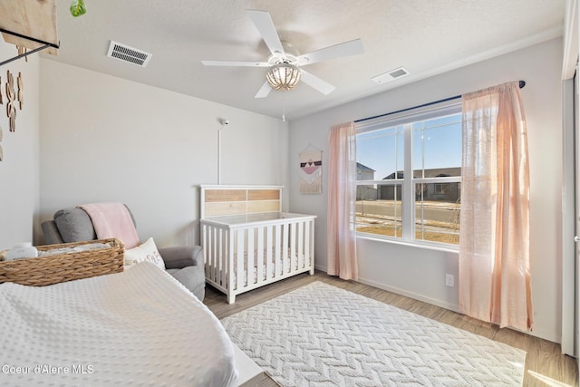 bedroom featuring a nursery area, ceiling fan, a textured ceiling, and light wood-type flooring