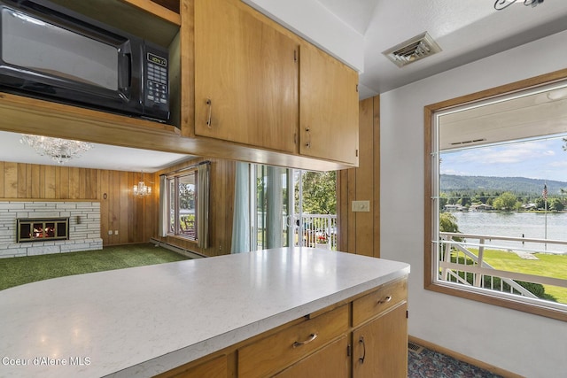 kitchen with a stone fireplace, a water and mountain view, dark colored carpet, and wood walls