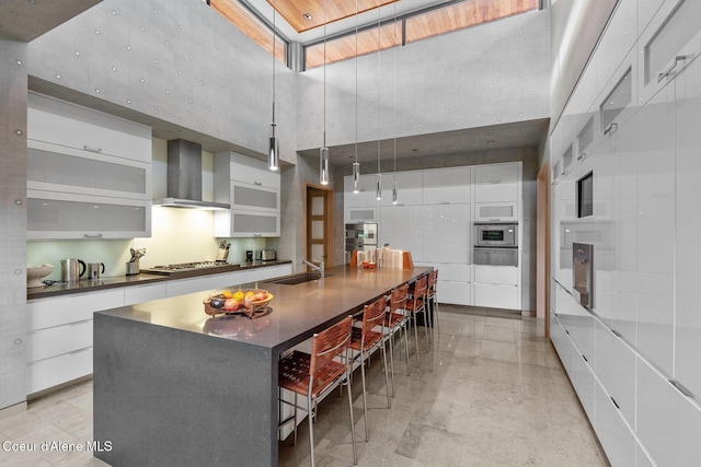 kitchen with a large island, wall chimney range hood, appliances with stainless steel finishes, white cabinetry, and a towering ceiling