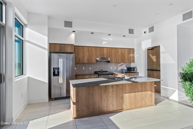 kitchen with sink, light tile patterned floors, appliances with stainless steel finishes, an island with sink, and decorative backsplash