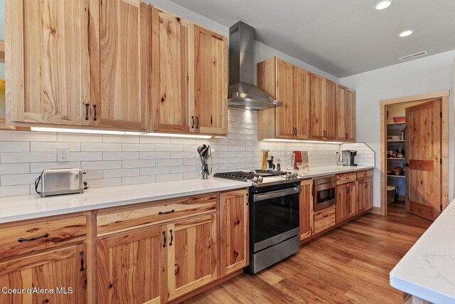 kitchen featuring appliances with stainless steel finishes, decorative backsplash, light stone counters, light hardwood / wood-style floors, and wall chimney exhaust hood