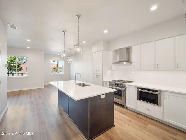 kitchen with wall chimney range hood, sink, white cabinetry, stainless steel appliances, and an island with sink