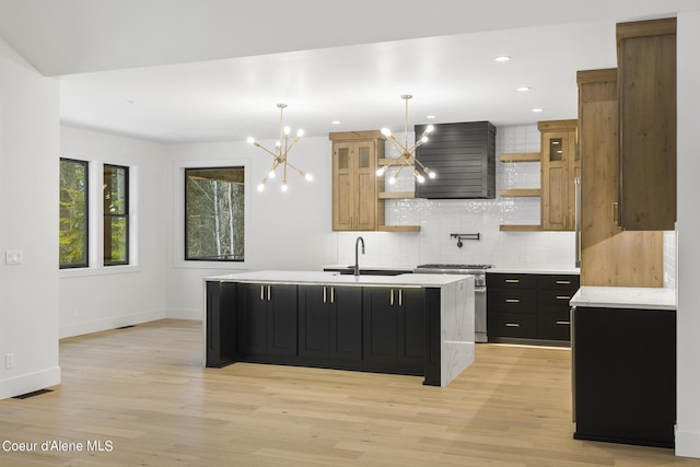 kitchen featuring decorative light fixtures, light hardwood / wood-style flooring, stainless steel range, a kitchen island with sink, and wall chimney range hood