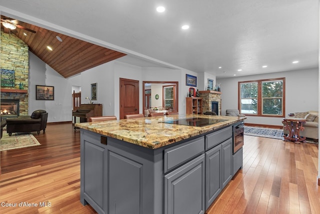 kitchen with gray cabinets, a stone fireplace, a kitchen island with sink, and black electric stovetop