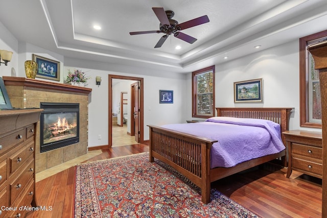 bedroom with a tiled fireplace, ceiling fan, a raised ceiling, and light hardwood / wood-style floors